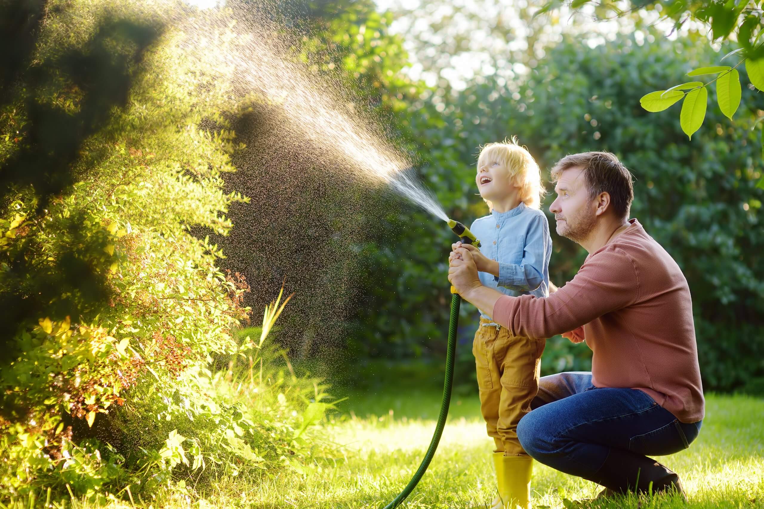 Funny Little Boy With His Father Watering Plants And Playing Wit