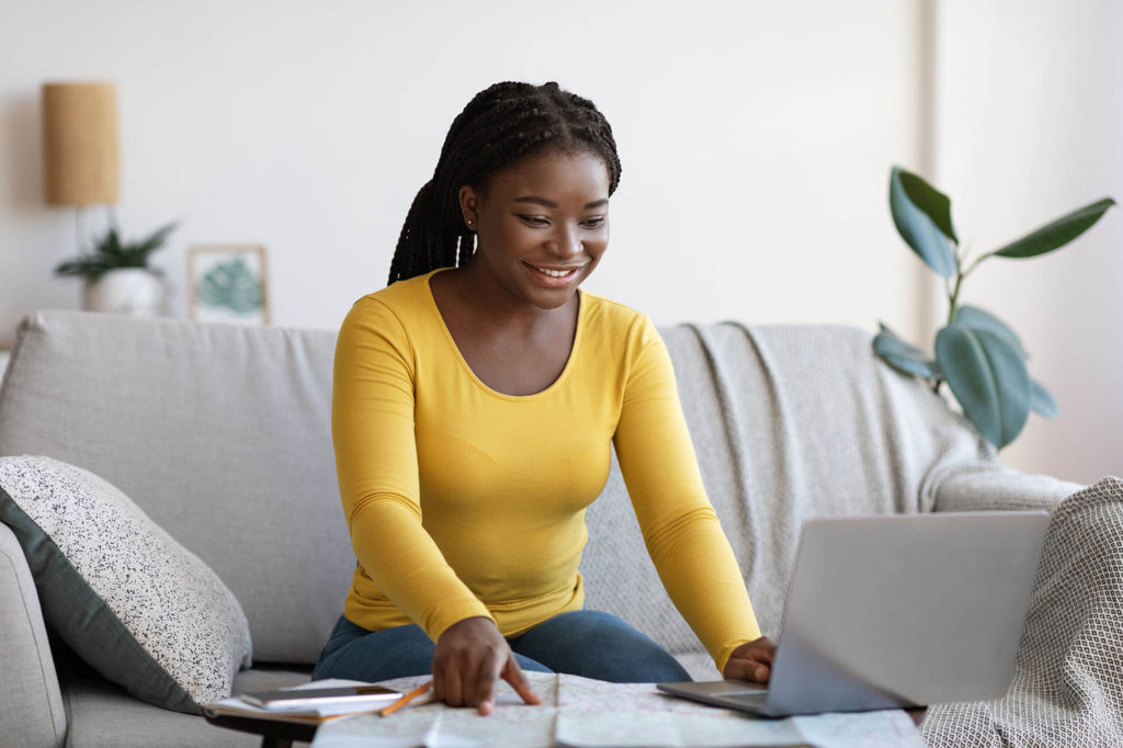 Smiling Black Lady Using Laptop And Map At Home, Planning Future
